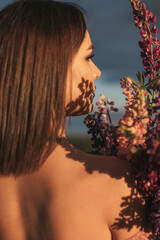 Close-up of a young woman with bare back and shoulders holding a bouquet of lupines in the sunset...
