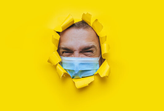 A young man in a protective medical mask looks in surprise and angrily with curiosity into a broken hole in yellow paper. The concept of pandemic, quarantine, protection against coronavirus, covid-19.