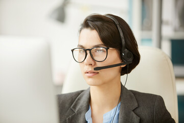 Serious pensive attractive hotline specialist in headset using video conferencing app while...