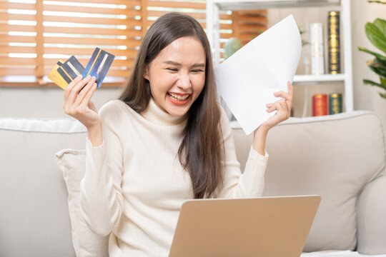 Happy Excited, Asian Young Woman, Girl Holding Credit Card And Paperwork Getting, Received Job Promotion, Approve Tax Refund Cash Back, Looking Good News At Laptop Computer, Sitting On Sofa At Home.