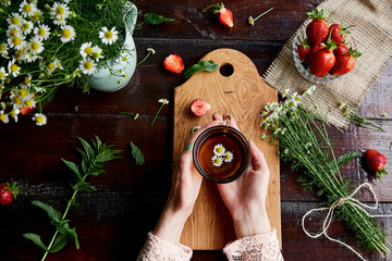 Woman hands holding cup of chamomile tea. Summer atmospheric table with fresh strawberries,...