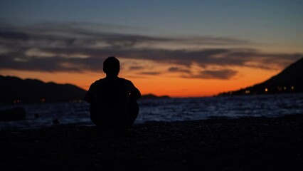 Young male sitting alone on a sandy beach watching a beautiful orange sunset over the tropical ocean in Croatia, Europe. - Powered by Adobe