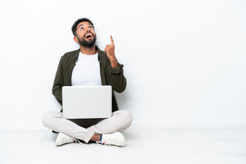 Young Brazilian man with a laptop sitting on the floor isolated on white pointing up and surprised