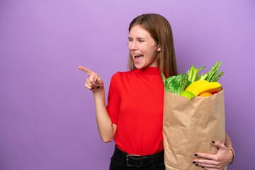 Young English woman holding a grocery shopping bag isolated on purple background pointing finger to the side and presenting a product