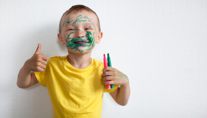 A 4-year-old boy painted his face with a marker. The boy stands against a white wall, holding markers in his hands, smiling and showing the class. Banner with space for text