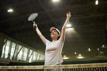 Fototapeta na wymiar Cheerful excited young woman in cap standing by net and raising arms up while celebrating winning in tennis match