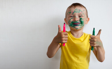 A 4-year-old boy painted his face with a marker. The boy stands against a white wall, holding...