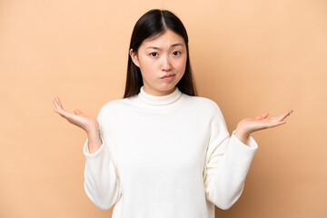 Young Chinese woman isolated on beige background having doubts while raising hands