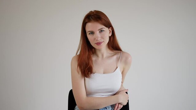 Beautiful red-haired young attractive woman sits on a chair and folded her hands on her knees, looking affably into the camera.