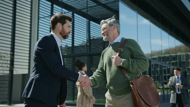 Two handsome Caucasian men handshaking, talking and gesturing outdoor. Meeting of colleagues on background of glass business center. Business people. Morning time. Beautiful modern architecture
