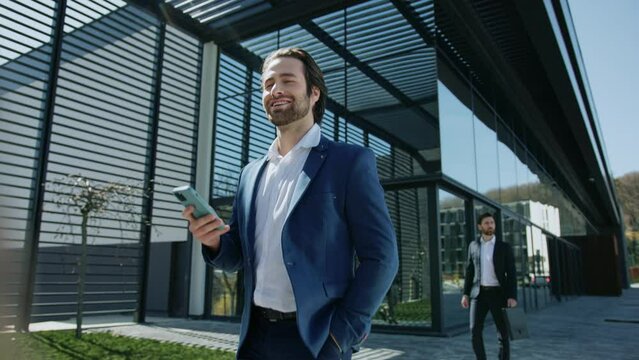 Footage of happy young smart man dressed in blue jacket walking near business center. Good-looking bearded Caucasian businessman holding smartphone in hand, looking at screen and smiling. Success