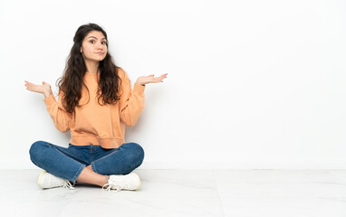Teenager Russian girl sitting on the floor having doubts while raising hands