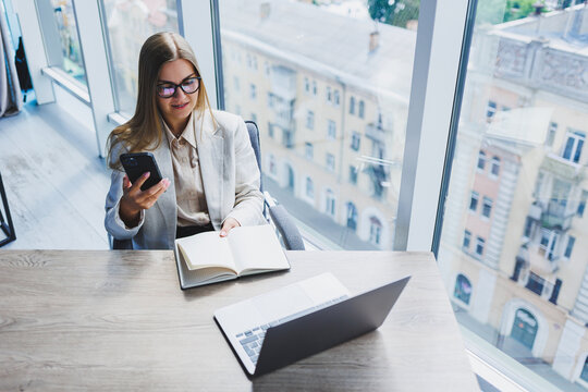 A Fair-haired Cheerful European Woman In Glasses In Stylish Casual Clothes Is Sitting At A Table With A Laptop, Doing Paperwork And Talking On The Phone. Business Lady At The Workplace In The Office