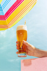 Human hand holding glass of cold light beer over summer blue sky background. Vacation, happiness,...