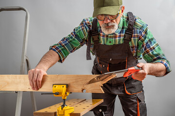 Aged worker working with a vise screwed to sawhorse. Senior man in overalls, cap and goggles