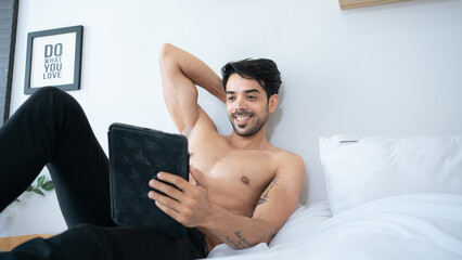 Muscular Hispanic man lying in bed under a black blanket with smartphone in hands. Concept of...