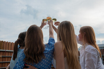 Young girls standing together, raising hands and clinking glasses of white wine closeup back view, sky background. Having fun and enjoying outdoor recreation. Girls party, celebration, friendship