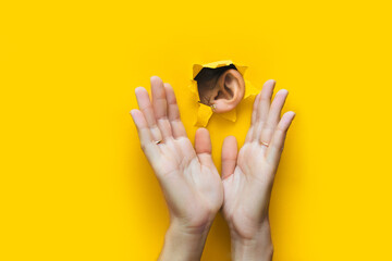 Close-up of a woman's ear and two hands through a torn hole in the paper. Yellow background, copy...