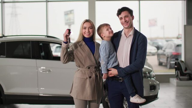 auto business, smiling man and woman with child holding keys to new vehicle while standing in a car center, looking at camera
