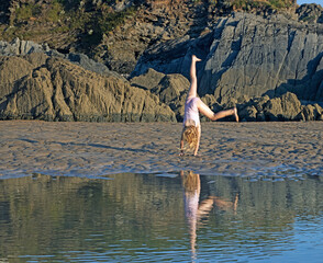 Female child (6-7 years old) performing acrobatics along sandy beach in North Devon reflected in...