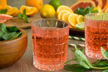 Two glasses full fresh natural lemonade, cocktail, infused water standing on wooden table. Slices of orange, grapefruits, lemon, lime, tangerine, spearmint leaves, wooden utensil on background