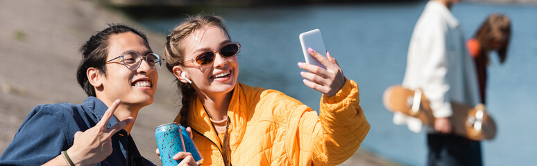 happy woman in sunglasses taking selfie with asian friend showing victory gesture, banner.