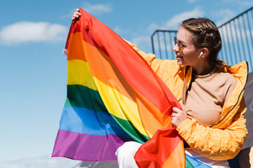 smiling woman in earphone holding lgbt pride flag outdoors.