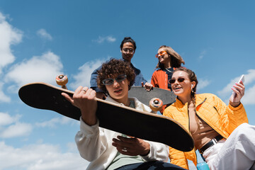 low angle view of young man holding skateboard near happy interracial friends outdoors.