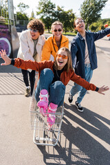 thrilled woman with outstretched hands riding in shopping trolley near interracial friends.