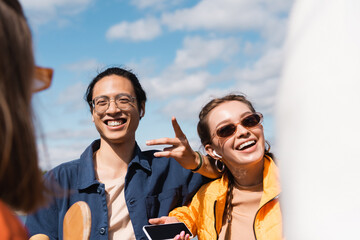 cheerful asian man pointing with finger near smiling woman in stylish sunglasses.