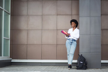 happy dark-skinned student girl with backpack and textbooks on background of university or college. concept of study. woman looks to left. copy space