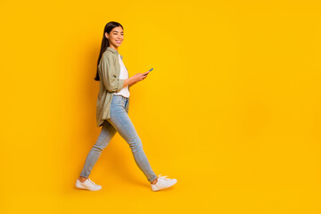 Fototapeta na wymiar Full length profile photo of pretty positive girl hold telephone walk empty space isolated on yellow color background