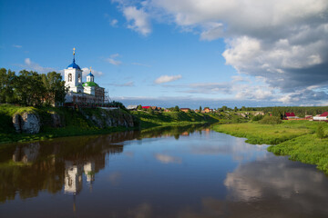 Obraz premium an old Orthodox church and a village on the river bank.sun , blue sky and black clouds.