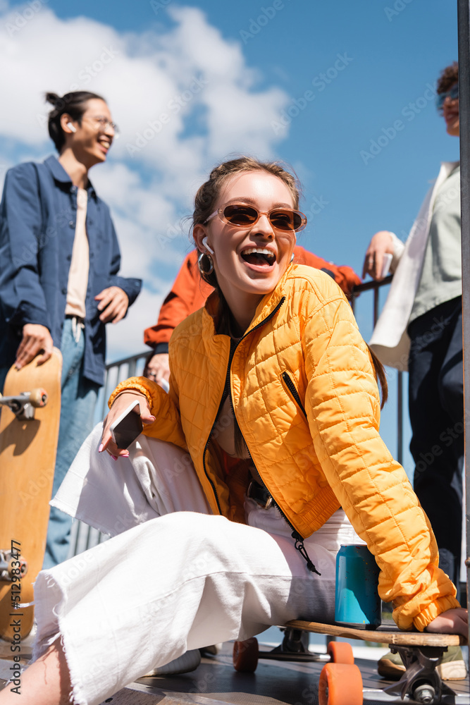 Wall mural amazed woman in sunglasses sitting near blurred multiethnic skaters against blue sky.