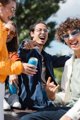 excited asian man in eyeglasses pointing with finger near blurred friends outdoors.