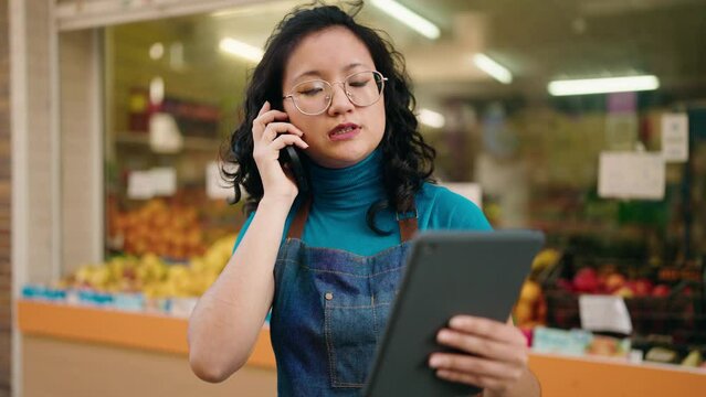 Young chinese woman employee talking on the smartphone using touchpad at fruit store