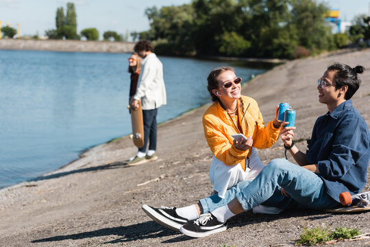 asian man sitting on longboard near river and clinking soda cans with cheerful friend.