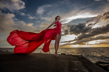 Beautiful lady in long red dress on the windy beach at the sunset.