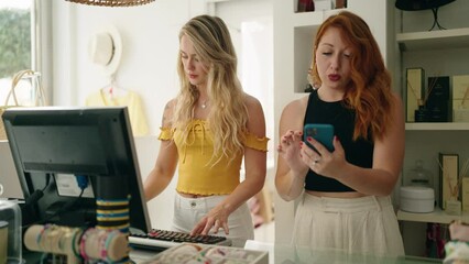 Two women shop employees using smartphone and computer working at clothing store