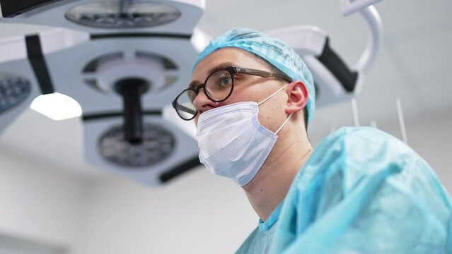 Young male doctor in medical mask, cap and glasses. Portrait of healthcare professional focused on work. Low angle view.