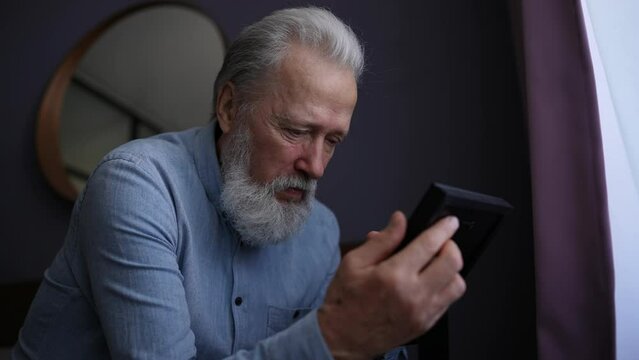 Close-up low-angle view of depressed elderly bearded man sitting on sofa at home in evening, looking at picture of wife in frame and crying. Concept of nostalgia, grief, longing, loneliness in old age