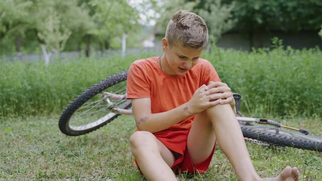 The young handsome boy is crying and upset that he fell off the bike and got painful leg injury. The child is holding the knee because the wound on the skin hurts. Accident with bicycle in green park