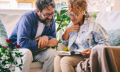 Young mature couple reading together a memories book diary outside home in the terrace sitting on couch. Happy man and woman leisure activity. People enjoying life and love. Caucasians with notes