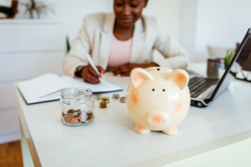 Closeup shot of coins and a piggybank on a table with an unrecognisable businesswoman working in...