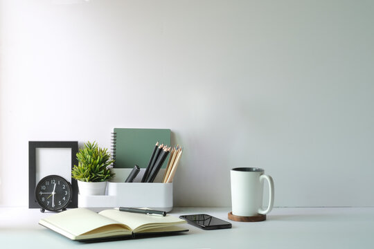 Home office desk with alarm clock, coffee cup, books, pencil holder and flower pot on white table. Copy space..