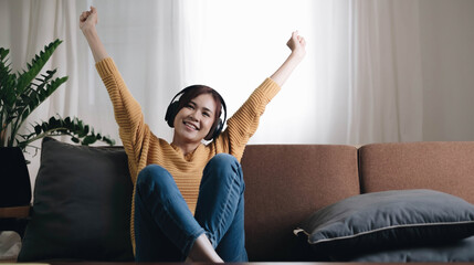 Being energetic. Pretty overjoyed young  woman laughing and listening to music and wearing headphones while sit on the sofa and wearing a yellow sweater