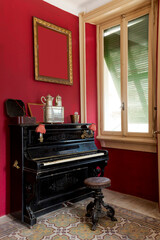 Detail of black piano with stool and shuttered window. There is a large frame hanging on the wall