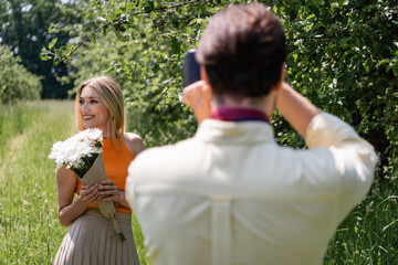 Obraz premium Positive woman with bouquet posing near blurred boyfriend with retro camera in park.
