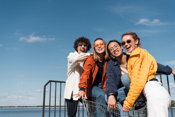 excited multicultural friends with shopping trolley laughing at camera against blue sky.
