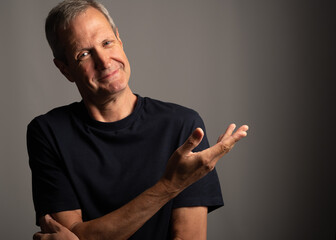 Smiling older man in a blue tee shirt, gesturing with his hand, on a grey background with copy space